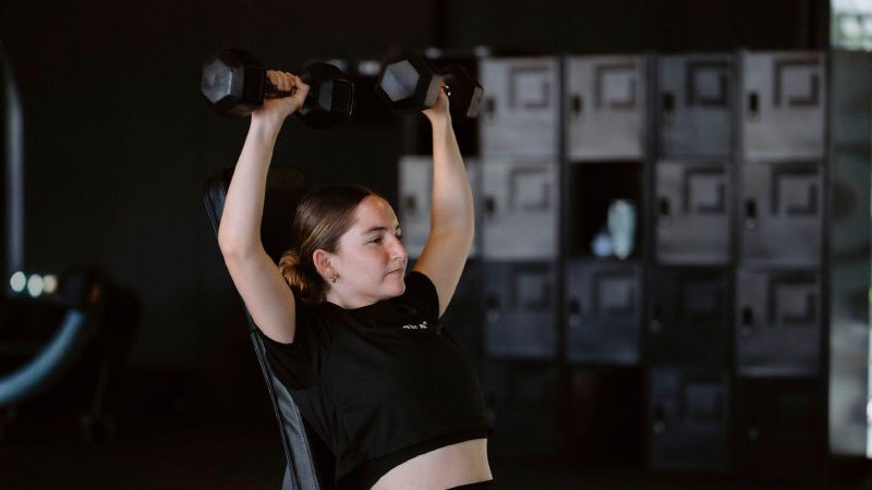 Een vrouw drukt twee halters boven haar hoofd in een sportschool