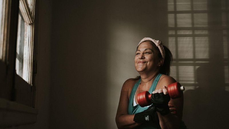 Een vrouw van middelbare leeftijd traint lachend met een halter bij het raam